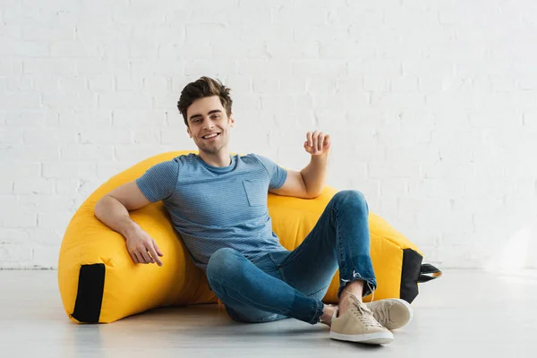 Glücklicher und gutaussehender Mann sitzt mit überkreuzten Beinen in der Nähe des gelben Bohnensackstuhls zu Hause — Stockfoto