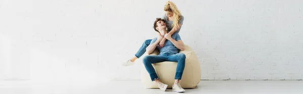 Panoramic shot of cheerful blonde girl hugging happy boyfriend sitting on beige bean bag chair at home — Stock Photo