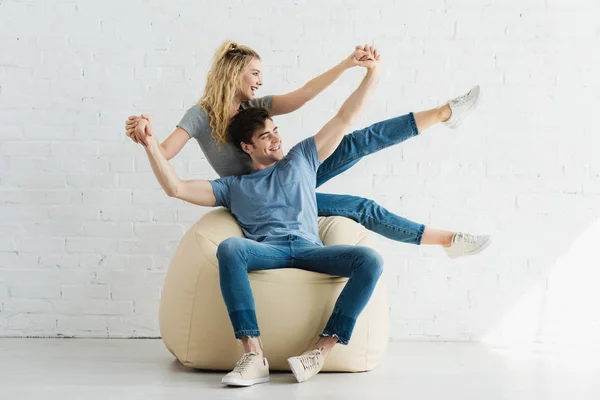 Cheerful blonde girl and happy man holding hands while sitting on bean bag chair — Stock Photo