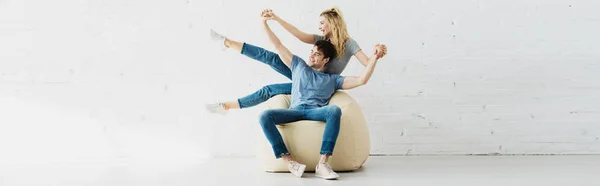 Plan panoramique de gaie fille blonde et homme heureux tenant la main tout en étant assis sur une chaise de sac de haricot — Photo de stock