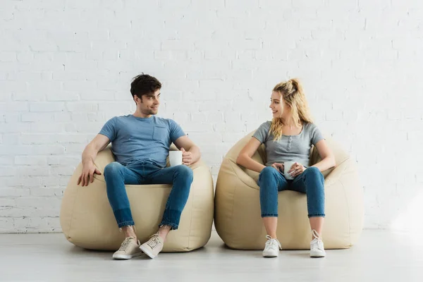 Glückliches blondes Mädchen und fröhlicher Mann, der Tassen hält, während er auf Sitzsäcken sitzt — Stockfoto