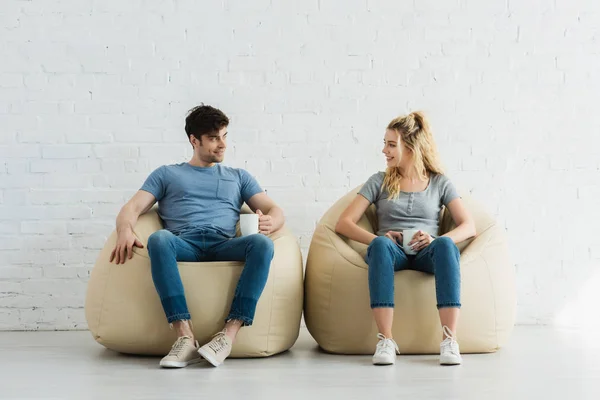 Feliz rubia chica y alegre hombre sosteniendo tazas mientras está sentado en frijol bolsa sillas - foto de stock