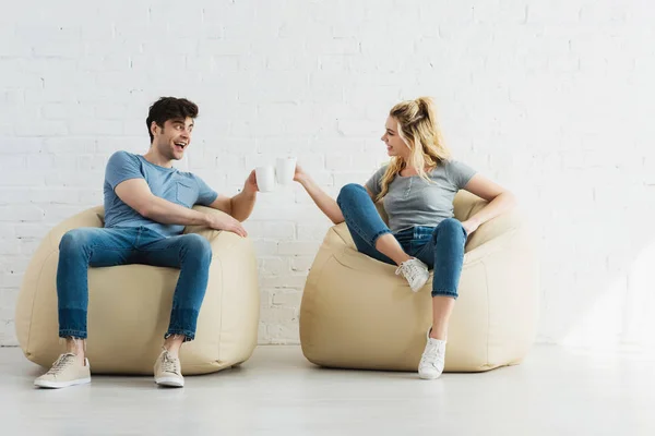 Attraktives blondes Mädchen und fröhlicher Mann, der Tassen hält, während er auf Sitzsäcken sitzt — Stockfoto