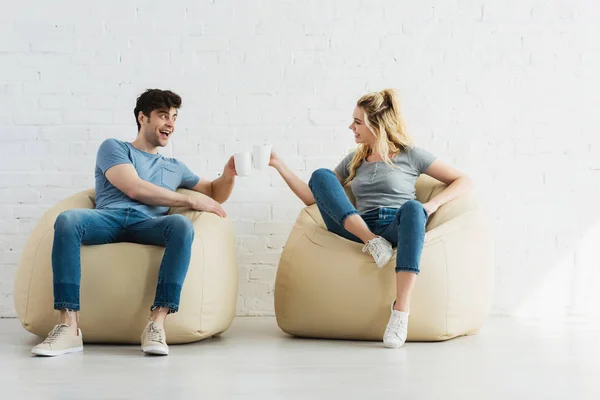 Atractiva chica rubia y hombre alegre sosteniendo tazas mientras está sentado en sillas de bolsa de frijol - foto de stock