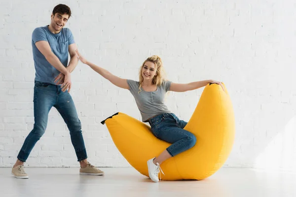 Cheerful woman sitting on yellow bean bag chair and touching handsome man — Stock Photo
