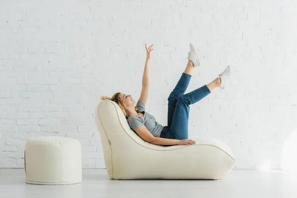 Cheerful blonde girl lying on  bean bag chair and gesturing near brick wall — Stock Photo