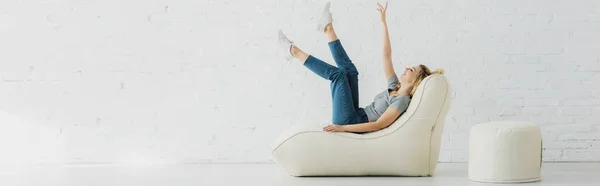 Plan panoramique de gaie fille blonde allongée sur une chaise de sac de haricot et geste près du mur de briques — Photo de stock