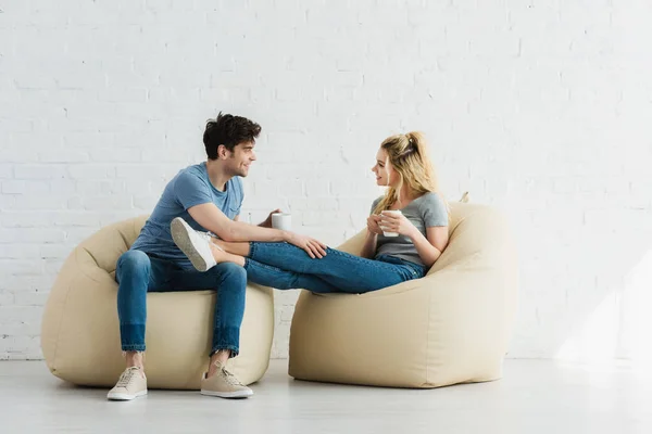 Cheerful woman and happy man sitting on bean bag chairs and holding cups — Stock Photo