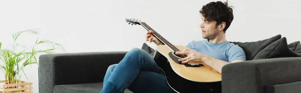 Panoramic shot of handsome man sitting on sofa and playing acoustic guitar at home — Stock Photo