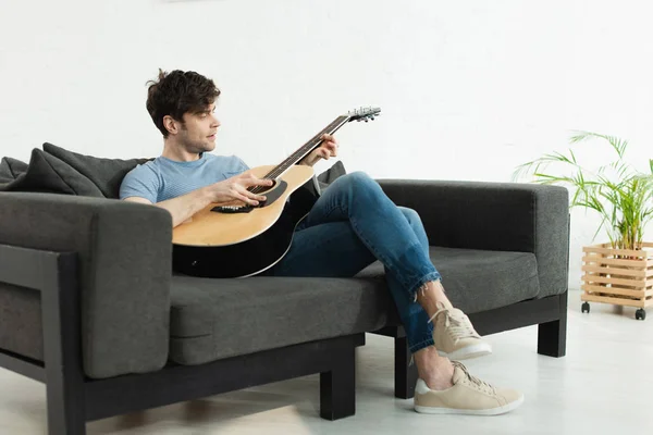 Hombre guapo sentado en el sofá y tocando la guitarra acústica en casa - foto de stock
