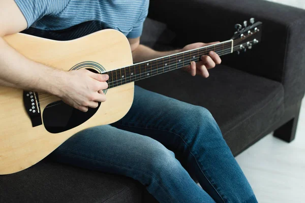 Vue recadrée de l'homme assis sur le canapé et jouant de la guitare acoustique à la maison — Photo de stock