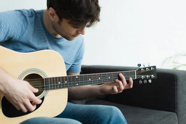 Guapo músico tocando la guitarra acústica mientras está sentado en el sofá en casa - foto de stock