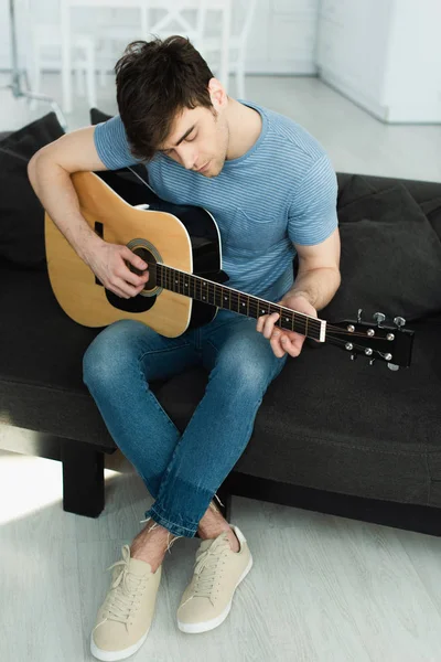 Músico bonito tocando guitarra acústica enquanto sentado com pernas cruzadas no sofá — Fotografia de Stock