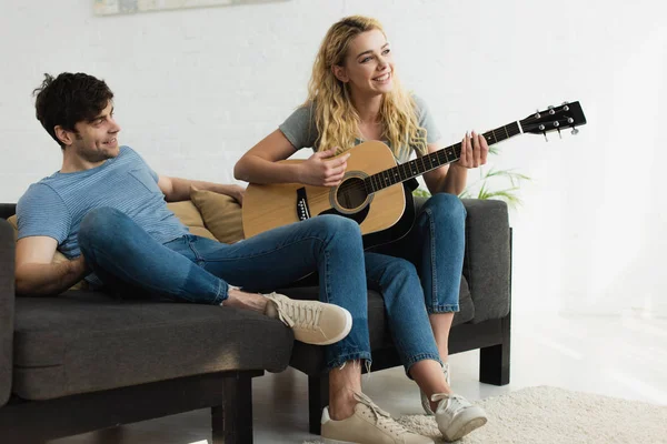 Glückliches blondes Mädchen spielt Akustikgitarre in der Nähe eines fröhlichen Mannes — Stockfoto