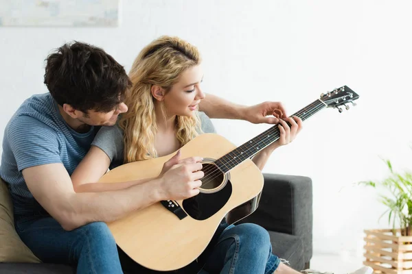 Красивий чоловік навчає блондинку щасливу жінку грати на акустичній гітарі вдома — стокове фото