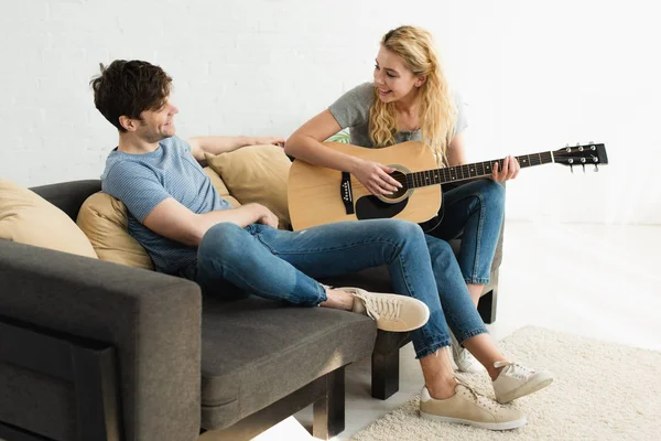 Chica rubia feliz tocando la guitarra acústica y mirando al hombre alegre - foto de stock