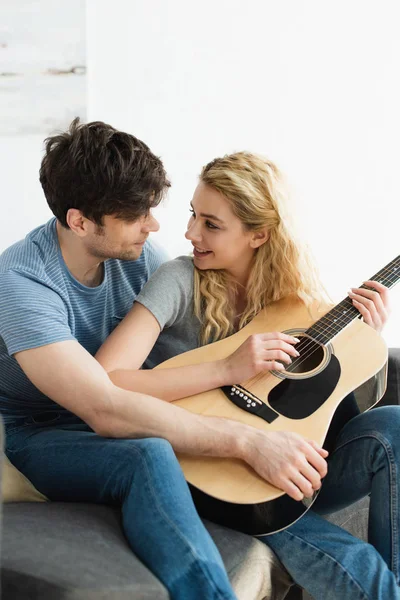Feliz rubia joven sosteniendo la guitarra acústica cerca de hombre alegre sentado en sofá - foto de stock