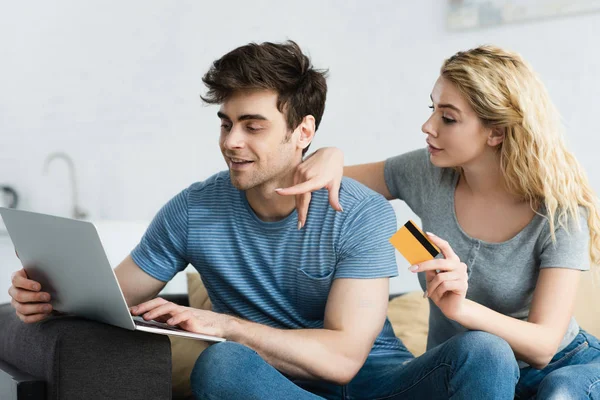 Mujer rubia sosteniendo la tarjeta de crédito y apuntando con el dedo a la computadora portátil cerca de hombre alegre - foto de stock