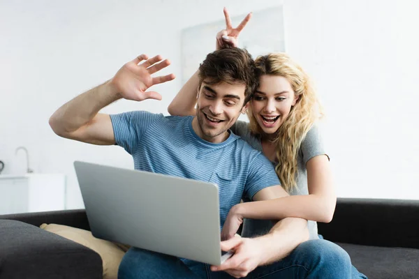 Cheerful man waving hand near happy woman showing peace sign while having video call on laptop — Stock Photo