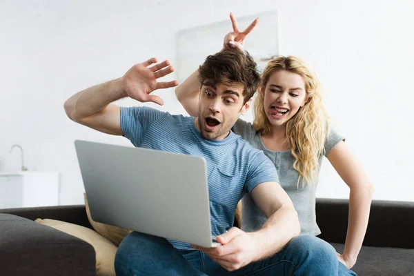 Веселый мужчина машет рукой возле счастливой женщины показывая язык и знак мира во время видеозвонка на ноутбуке — стоковое фото