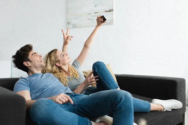 Fröhlicher Mann zeigt Friedenszeichen und blondes Mädchen macht Selfie zu Hause — Stockfoto