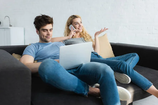 Allegra ragazza bionda che parla su smartphone vicino all'uomo usando il computer portatile — Foto stock