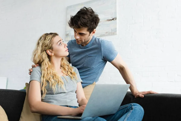 Hombre guapo mirando atractiva chica rubia sentada con el ordenador portátil - foto de stock