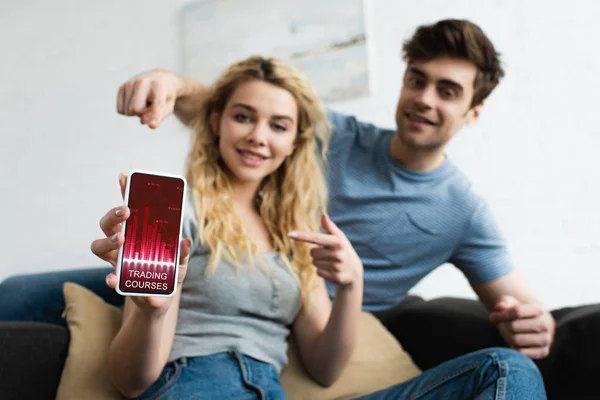 Selective focus of cheerful man and blonde woman pointing with fingers at smartphone with trading courses app on screen — Stock Photo