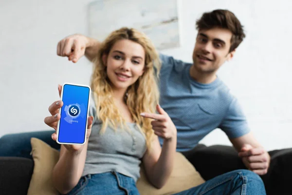 Селективний фокус веселої людини і білявої жінки, що вказують пальцями на смартфон з додатком Shazam на екрані — стокове фото