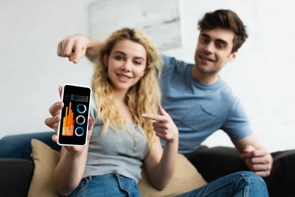Вибірковий фокус веселого чоловіка та блондинки, що вказує пальцями на смартфон з графіками та графіками на екрані — стокове фото