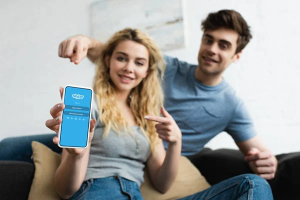 Селективний фокус веселої людини і білявої жінки, що вказують пальцями на смартфон з додатком Skype на екрані — стокове фото