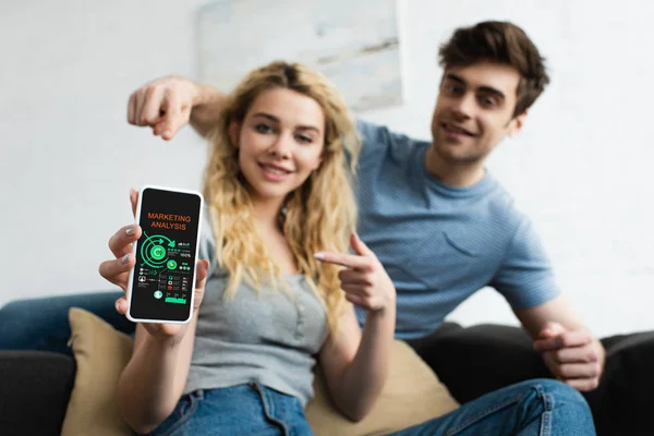 Selektiver Fokus glücklicher Männer und blonder Frauen, die mit den Fingern auf das Smartphone zeigen, mit Marketing-Analyse-Schriftzug auf dem Bildschirm — Stockfoto
