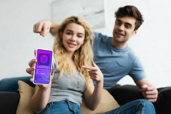 Избирательный фокус веселого мужчины и блондинки, указывающие пальцами на смартфон с приложением для покупок на экране — стоковое фото