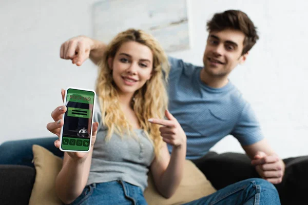 Избирательный фокус веселого мужчины и блондинки, указывающие пальцами на смартфон с бронированием приложения на экране — стоковое фото