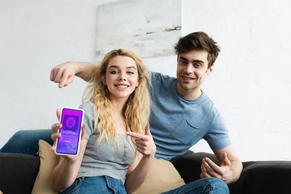 Избирательный фокус счастливого мужчины и блондинки, указывающие пальцами на смартфон с приложением для покупок на экране — стоковое фото