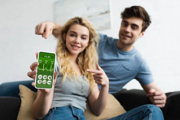Избирательный фокус веселого мужчины и блондинки, указывающие пальцами на смартфон с приложением для здоровья на экране — стоковое фото