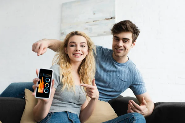 Messa a fuoco selettiva di uomo felice e donna allegra che punta con le dita a smartphone con grafici e grafici sullo schermo — Foto stock