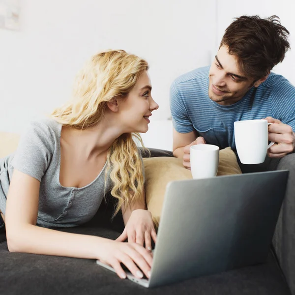 Homem feliz e bonito segurando copos perto de menina loira com laptop — Fotografia de Stock