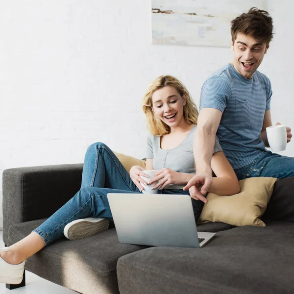 Uomo felice che tiene la tazza e punta con il dito al computer portatile vicino allegra ragazza bionda seduta sul divano — Foto stock