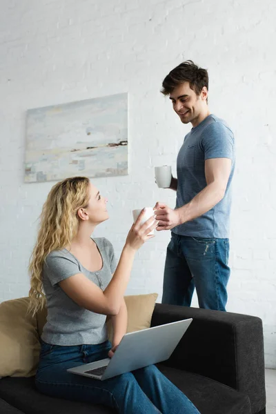 Homme heureux donnant tasse de café à gaie fille blonde assise sur le canapé avec ordinateur portable — Photo de stock