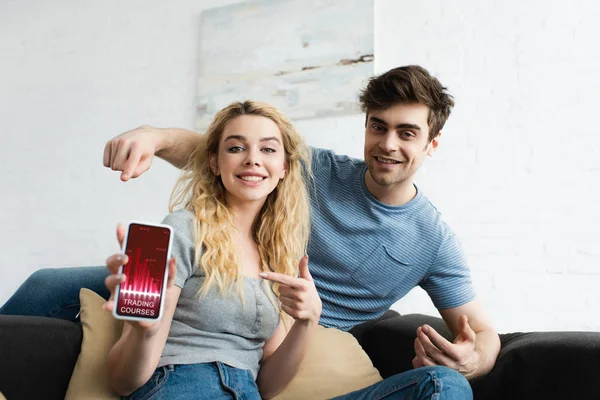 Messa a fuoco selettiva di uomo felice e donna bionda che punta con le dita verso smartphone con corsi di trading app sullo schermo — Foto stock
