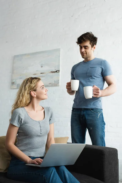 Hombre feliz sosteniendo tazas de café cerca de chica rubia alegre sentado en el sofá con el ordenador portátil - foto de stock