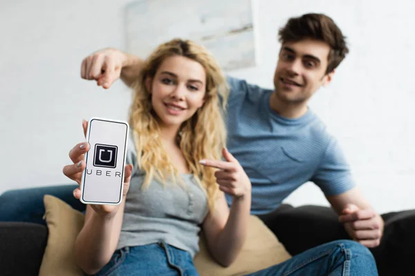 Селективний фокус веселої людини і білявої жінки, що вказують пальцями на смартфон з додатком Uber на екрані — стокове фото