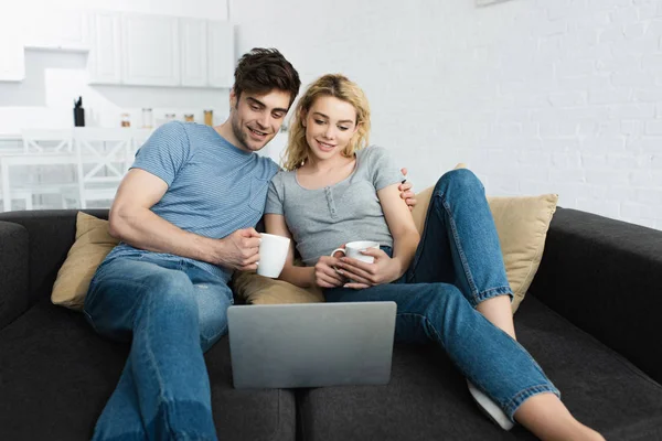 Hombre feliz sosteniendo la taza y viendo la película con chica atractiva en el ordenador portátil - foto de stock
