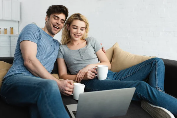 Hombre feliz sosteniendo la taza y viendo la película con chica alegre en el ordenador portátil - foto de stock