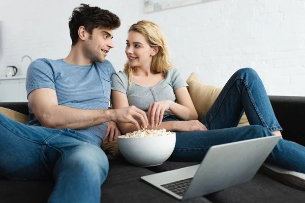 Happy man and cheerful blonde woman taking popcorn from bowl near laptop — Stock Photo