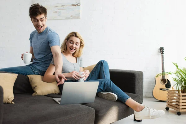 Schöner Mann zeigt mit dem Finger auf Laptop, während er Tasse in der Nähe attraktive blonde Mädchen hält — Stockfoto