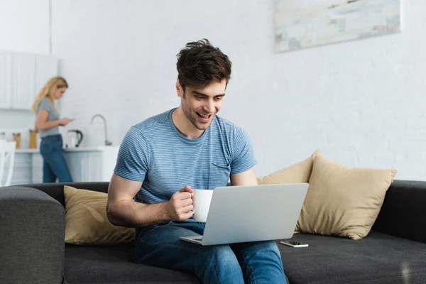 Selektiver Fokus des fröhlichen Mannes mit Tasse und Blick auf Laptop in der Nähe der Frau — Stockfoto