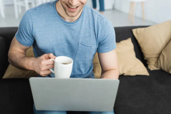 Vista cortada de homem feliz usando laptop enquanto segurando copo com bebida — Fotografia de Stock