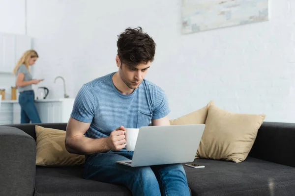 Messa a fuoco selettiva di bell'uomo che utilizza il computer portatile mentre tiene in mano tazza con bevanda — Foto stock